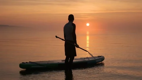 Man standing in water with stand up paddle board against background of sunrise. Video stock 244603635