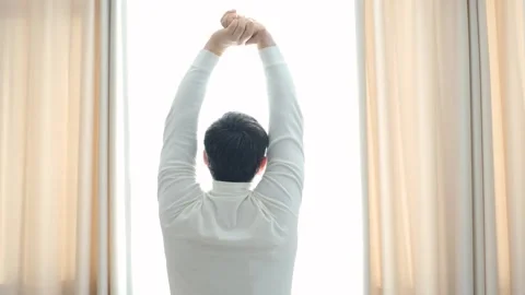 Man standing by window raising hands up and stretching, concept of early Stock Footage 301025084