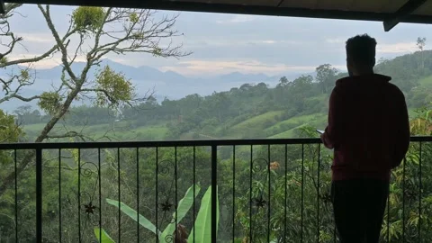 Man stands on a covered balcony in rainy mountain landscape. 動画素材 330085597