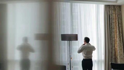A man stands facing a large window and gets dressed going to a meeting 스톡 동영상 168524253