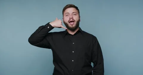 A man stands in front of a plain blue backdrop, making a phone gesture with.. Stock-Footage 326798797