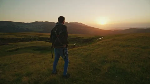 Man stands on grassy hill watching sunset over mountains in the background with Stock Footage 345640339