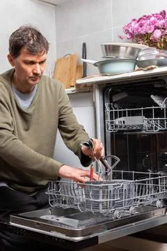 A man stands in his kitchen taking clean dishes out of the dishwasher. He foc Stock Photos
