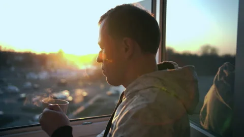 A man stands in an open window watching the sunset and drinking hot tea Stock Footage 130539279