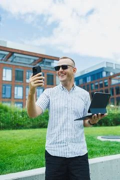Man stands outside in a modern setting holding a laptop and a smartphone wh.. Stock Photos
