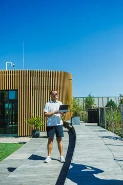 Man stands on a path holding a tablet outside modern building on a clear day Stock Photos