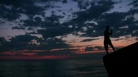 Man stands on rock and throws pebbles into sea under sunset sky Stock Footage 673469