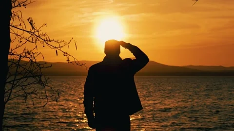 A man stands on the shore of a lake at sunset and looks into the distance. Vídeo Stock 337563700