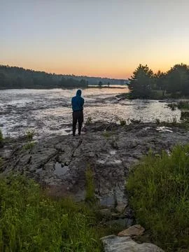 A man stands on the side of a river while admiring the sunset and forest Stock Photos