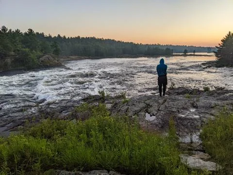 A man stands on the side of a river while admiring the sunset and forest Stock Photos