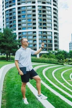 Man stands on steps in park, looking at phone, surrounded by green grass an.. Stock Photos