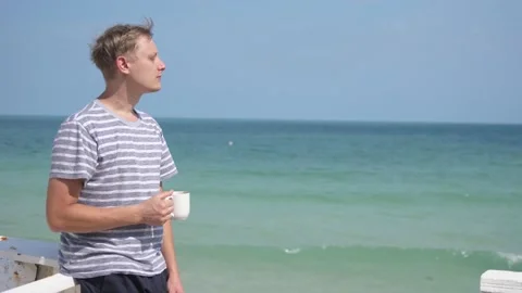 Man stands by the tropical beach and drink hot coffee from white mug.slow motion Stock Footage 200072405