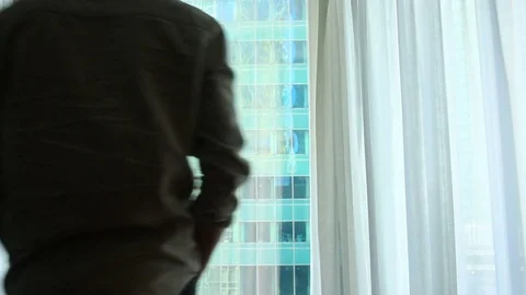 A man stands by the window with curtains in a hotel room, looks at an amazing Stock Footage 76497574