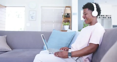Man is starting work, typing on laptop while wearing headphones on gray couch in Stock Footage 340981827