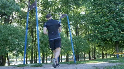 Man starts doing pull-up exercising on fitness loops in summer park. 4K slow Stock Footage 98796306