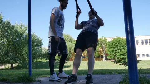 Man starts doing pull-ups on fitness loops under control of bearded fitness Stock Footage 99496251