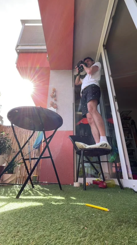 Man stepping down from chair on balcony while holding camera under bright Stock Footage 344883759