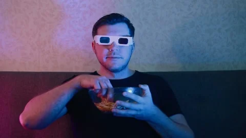 A man in stereo glasses sits on a sofa eating chips from a stained plate. Stock Footage 131440852