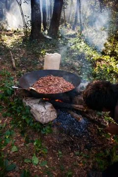 Man stirring up fire while cooking meat in wadjan on top of a stone in forest. Stock Photos