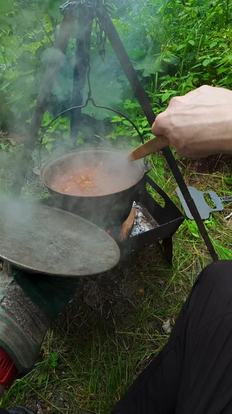 Man stirring steaming stew in campfire cauldron for outdoor picnic cooking .. Video stock 346146061