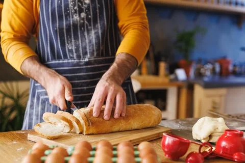 Man in striped apron slicing bread on board at kitchen counter with eggs and Stock Photos