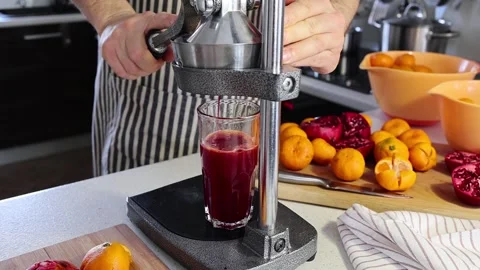 Man in a striped dark apron squeezes juice from pomegranates and tangerines Stock Footage 169343820