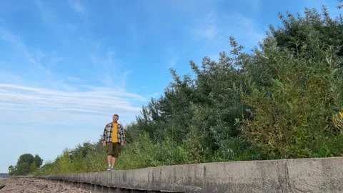 A man strolls along a concrete path bordered by lush greenery, set against a Stock Footage 292771716