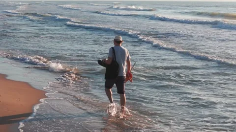 A man strolls on the beach at sunrise, carrying an orange umbrella and a bl.. Video stock 283973366