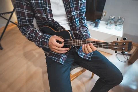 Man strums a black ukulele while seated indoors, focusing intently on his mus Foto stock