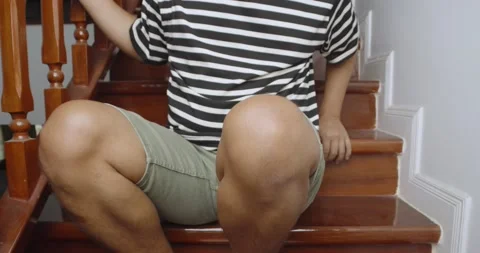 Man suffering from knee pain while sitting on the stairs at home. Stock Footage 238930807