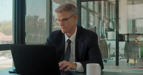 A man in a suit and glasses working on a laptop at the office with big window Stock Footage 140182098