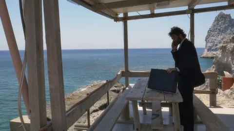 Man in suit with briefcase walking towards a wooden balcony while talking on his Stock Footage 77743407