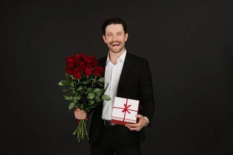 Man in suit is posing on black background with gift box and red roses in hands 스톡 사진