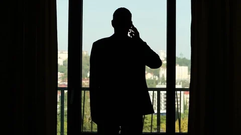Man in suit silhouetted stand against room window, answer on call Stock Footage 80741622