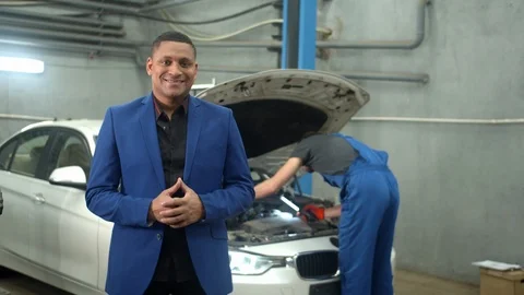 Man in suit smiles at the camera, mechanic repairs a car on the background Video stock 124254902