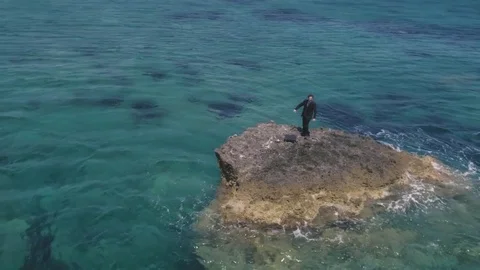 Man in suit stranded on a deserted rock in the middle of the ocean waving his Stock Footage 77922480