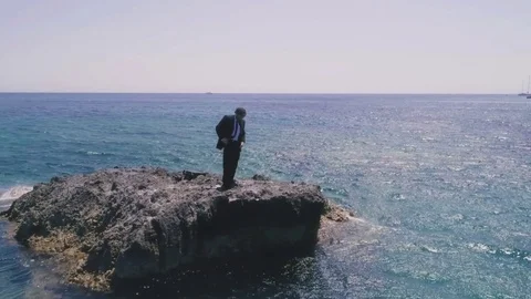 Man in suit stranded on a deserted rock in the middle of the ocean fixing up his Stock Footage 77926058