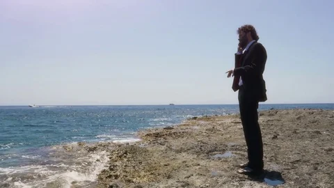 Man in suit talking on his smartphone on a deserted island in front of the ocean Stock Footage 77751289