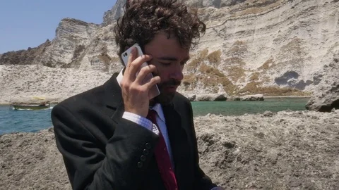 Man in suit talking on his smartphone on a deserted island in front of the ocean Stock Footage