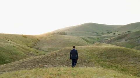 A man in a suit walks through the hills towards the sunlight. Spring green Stock Footage 275189741