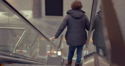 A man with a suitcase on wheels descends down an escalator in a building  Stock Footage 119596475
