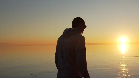 Man in sun protective glasses is on the beach along the sea at sunset Stock Footage 129639314