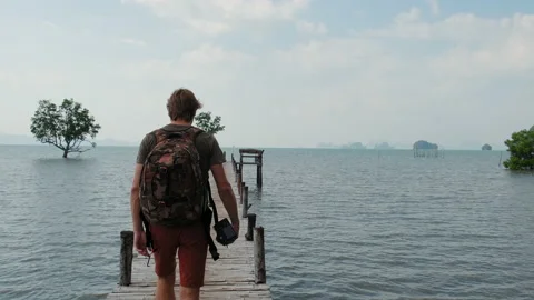 Man in sunglasses with backpack walks over a wooden bridge on the sea Stock Footage 130398239