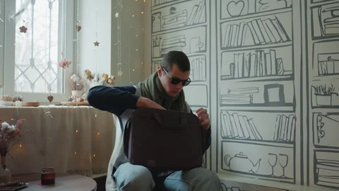Man in sunglasses opens laptop bag in cozy bookcase backdrop, morning light Stock Footage 345168513
