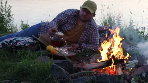 A man at sunset sits by the fire and the water and drinks tea Stock Footage 278384218