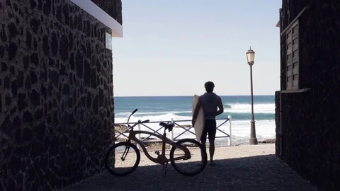 Man with surfboard standing between stone buildings and looking at the sea Stock Footage 71549383