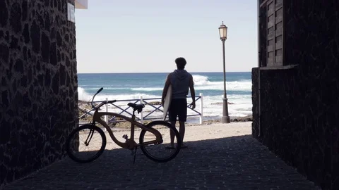 Man with surfboard standing between stone buildings and looking at the sea Stock Footage 71549650