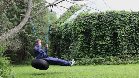 Man sways on swing which made from car tyre on tree at summer day. Slow motion Stock Footage 276376408