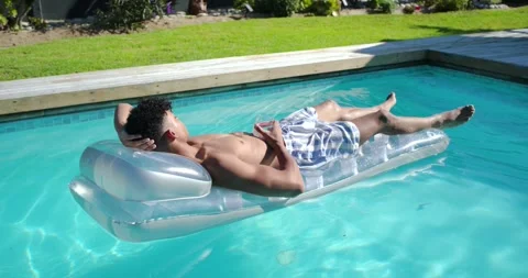 Man in swim-trunks lying on clear-float in pool with cell, camera pulling back Stock Footage 335747999