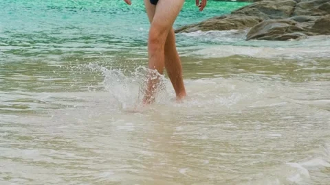 Man in swim trunks walking on sandy beach in shallow tropical sea water near Video stock 322070473